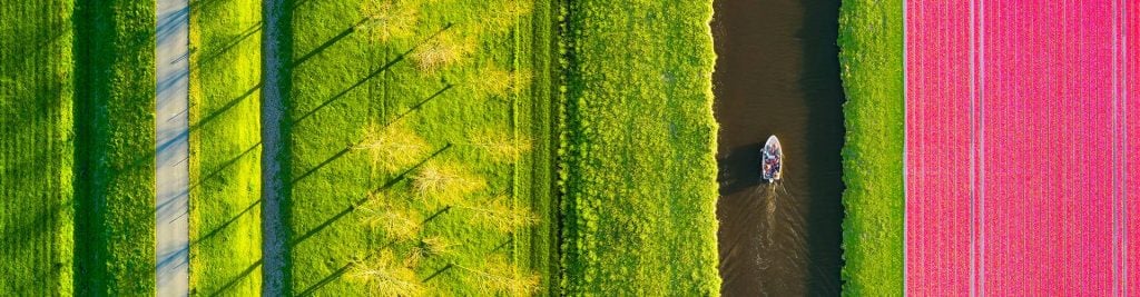 Aerial view of a small boat sailing along a river with lush, colourful fields on either side