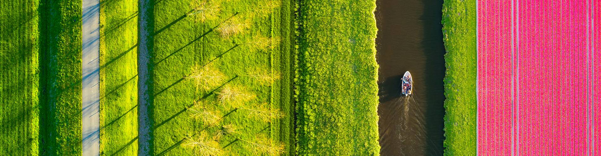 Aerial view of a small boat sailing along a river with lush, colourful fields on either side